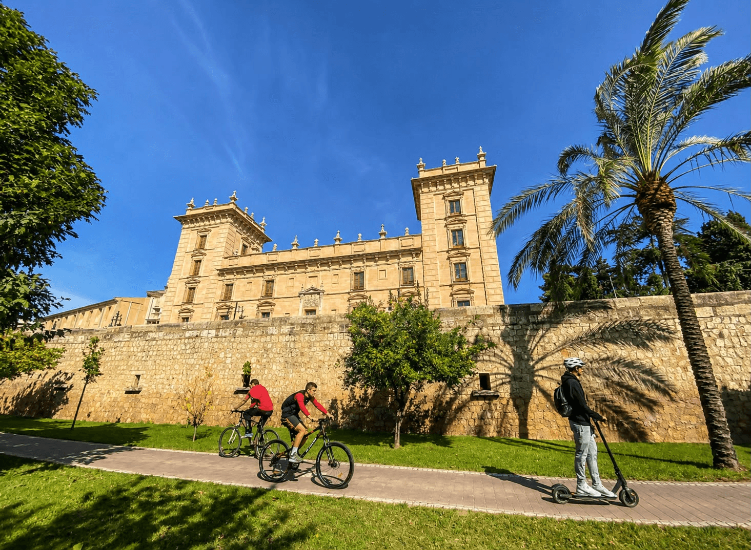 Ciclistas y una persona en patinete recorriendo los jardines junto al Museo de Bellas Artes de Valencia